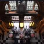 Looking down into a hallway from a great height. The far wall contains leaded windows in an arch-shaped frame, above wide doors with yellow lit panels around them. The floor has a geometric tiling pattern, and is seen beneath a simple chandelier.