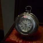 A large pocket-watch shaped clock, made of silver with a white face, on a red and gold cushion.