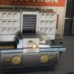 A small plastic toy, shaped like a 1960s computer. There is a grey panel with lights and buttons, and below a conveyor-belt like system with a crank handle. The cardboard box is behind, on which is visible “ELECTRONIC QUESTION AND ANSWER COMPUTER”, “the machine that thinks like a man”, “IT THINKS! IT ANSWER IT REMEMB” and “Feed it the question here”.