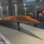 A wooden model of a streamlined aircraft, in a glass case. Portions of other models are visible, including a red and black ball on a stick. A gallery is (just) visible behind.
