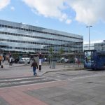 A station forecourt. Two roads for buses run across the centre, crossed by a zebra crossing; a bus in at one of the stands. The buildings around the forecourts are rectangular in profile, with mirrored panels interspersed with strips of windowpanes.