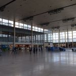 A high-roofed, large, interior space. The back wall is glazed, with shop fronts below. The left wall (the only other one visible) has a windowed frontage (though the windows are not to the outside), with a sign for “John Lewis”. The hall is mostly empty, with some people passing through, and a couple of cars.