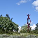 A sculpture that looks like human figures, with arms outstretched, forming a vertical chain, made of copper-coloured metal. There are shrubs and trees around, and the sculpture was set against a blue sky with small fluffy clouds.