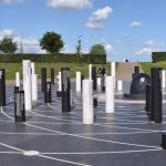 A tiled area with black tiles inset with various white orbit-like rings, on which are set white and black pillars of various heights, with inscriptions of dates and text. The centre of the circular paved area is a pine-cone-like shape, made of black stone, surrounded by rings of coloured paving. There are a few people around. In the background are grassy banks topped by hedges, and a path leading to a metal pyramid. The sky is bright blue with white fluffy clouds.