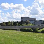 A grassy verge of a road in cutting, spanned by a late concrete bridge. Behind are trees and a concrete-and-glass building made of various units. The sky is bright, but cloudy.