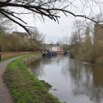 A canal enters the picture from the right and curves around, in a view taken from the near-empty towpath (two people appear in the background). A bare tree overhangs at the top. There is light, mostly brick, development around the canal.