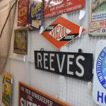 An array of advertising signs on a wide white metal mesh: e.g. “REEVES” “OFFICIAL VIGZOL STOCKIST”. Some netting hangs on the mesh behind. In the background is farm equipment behind glass.