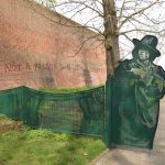 A green fence, partly shaped like theatre curtains and partly in a cut-out of Oscar Wilde, this latter part overhanging a path. Most of the fence is over grass, and behind it is a bare tree. To the left is a high brick wall, on which is spray-painted (black with pink accents): “I’M NOT A PRISON, darling, I’M A THEATRE”.