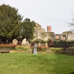 In the foreground, a grass patch surrounded by low walls and hedges, with a copper statue. Behind, a lush tree partially masks ruined stone walls, containing some arches, with a brick building with chimney behind.