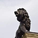 A bronze lion, with muscles detailed on its body, stands on a plinth (mostly out of shot) against a cloudy sky. The lion is roaring.