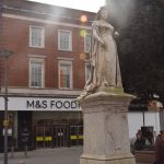 A yellow stone life-sized statue of queen Victoria on a plinth, in front of a three-storey brick shop building, the lower floor of which has signs reading “M&S FOODH[obscured]” and “COLLECT BY C[obscured]”.