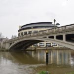 An arched metal bridge, painted white, with a balustrade at the top, over a wide muddy river. Across the river are office buildings.