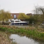 An area of marshland lies in front of a watercourse, with trees on either side. A motorboat is on the watercourse, and there are partly obscured buildings on the far side.