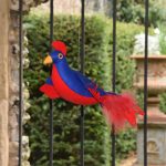 A stuffed toy bird with red and blue plumage is suspended by a wire. It is seen in front of a wall and gate, behind which, in soft focus, are hedges and an urn containing flowers.