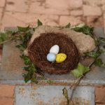 A nest and brightly-coloured small eggs are set on a rock and some bright green foliage, which is on a metal pillar against a red stone floor.