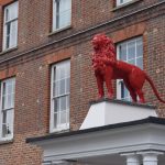 A painted red lion stands on a white-painted portico. The portico belongs to a brick building, with rows of windows with some Art Deco detailing above.