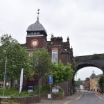 A street leading away from the camera, and passing under a blue brick railway arch. To the left of the arch is a dark brick building, three stories, with a squat clock tower.