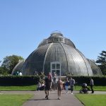 A dome-shaped greenhouse, viewed from one end, against trees and a bright blue sky. People walk along a path in front, with passes through a lawn in front of a hedge.