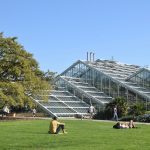A conservatory made up of several sections which are triangular prisms fused together. In front of it are some trees, small plants and a lawn; people sit on the lawn, and some walk past on a path.