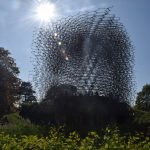 A metal structure in a cube shape, made up of metal rods forming a honeycomb shape. This sits against a bright blue sky, with a sun flare at the top. Around it is long grass, and there are bright blue trees behind. The cube sits on a mound, and there are people inside.