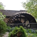 A water wheel, made of wood, attached to a low brick building with a dark, mossy roof. The wheel is set in a garden with blue flowers, surrounded by trees; in the distance is a field.
