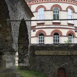 A ruined building. On the left are two stone arches, with the back wall being low, also of stone, with a small bricked-up rough fireplace. The whole structure is supported by metal poles. Behind is a yellow-brick building with red-brick and terracotta detailing; two storeys, with arched windows. The building has a protruding curved section was a flat wall to the left, and the inscription “SCHOLA REGIA GRAMMATICA MDLXII”.