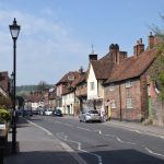 A street in a village with cars parked along both sides. The buildings are old, of brick, some painted white or half-timbered. There are gas-style lamp-posts along one side of the street.