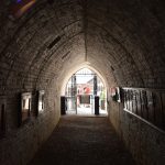 Looking along a vaulted brick-lined tunnel. The tunnel is dark, with display boards just visible on both sides. Light fours in from the end, which has a gate and turnstile, and three people can be seen standing in the outside.
