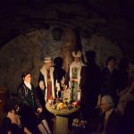 A group of waxworks, some standing and some seated, gather around a table on which are placed vessels, fruit and a candelabra. The two central waxworks wear fur hats and fur coats, and necklaces, in a form of regalia. The scene is in a cave, and fairly dimly lit.