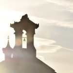 A stone archway containing a small bell, on the top of a hipped roof. There are some other turrets. The photo is taken with a sun flare behind the tower.