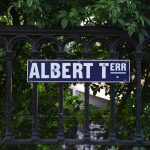 A street sign reading “ALBERT TERR.” in white on blue, against a black painted railing with arches between the metal posts. Behind the railing is dense foliage.