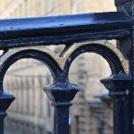 A close-up view of the top of railings, with arches between the metal posts. The railings are painted black, except for gold details on the arches. Behind, out-of-focus, are a light stone mill buildings, several storeys high.