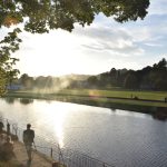 A view across a lightly rippled river towards parkland, with a sun flare in the sky. On the near bank is a paved area with people milling around, and a tree to the left in the foreground.