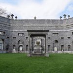 Inside a large octagonal structure, with grey stone walls, open to the elements (the ground is grassed over). In the centre stands a form of portico, with a tomb underneath, and there are several other memorials on the walls. The corners of the walls are topped by urns. The sky is blue, and trees are visible behind the walls.