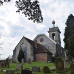 A church in a graveyard. The walls of the church are of grey stone, with red brick detailing. A concrete-esque tower is at the far end, with a green structure supported a (dirty) large golden ball. The sky is cloudy but bright.