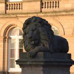 A stone lion appears dark on a plinth against the building behind, which is drenched in sunlight. The lion is reclining, and licking its paw, with an inscription on the plinth reading “PEACE”. The building behind has four high arched windows, and above four sets of stone railings for the storey above (out of shot).