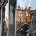 A pair of floor-to-ceiling shop windows either side of a door. Reflected in the nearer one are buildings on the other side of the street, including a three-storey building with ornate detailing. Reflections of further up the street are also visible in the other window.
