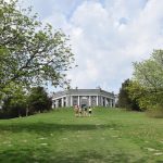 A view up a grassy hill, with an informal path up it with trees and bushes on either side. A group of four climb the hill ahead, with more people further up. At the top of the hill is an octagonal structure, of grey stone, with white columns and detailing, and high open arches (three on each side).