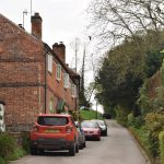A steep road, with old-fashioned lamposts, and cars parked along it. On the right is a high hedge, with a small garden of shrubs in front. On the left are houses, of brick, with green detailing around the white windows and porches. The houses are quite large but are attached.
