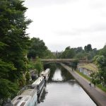 A view along a canal from a bridge, with a few people on the towpath. There is another bridge across the canal and, on the horizon, a spiky structure. There are thick trees on either side.