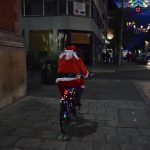A man dressed as Santa on a bike decorated with fairy lights rides down a street in a city centre at night. The street itself is decorated with Christmas lights.