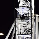 A view of a cabin of a floodlit Ferris wheel against the dark sky. Two people are in the cabin, silhouetted against the structure of the wheel.