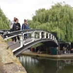 A simple arched metal footbridge, painted black-and-white, over a canal, viewed almost end-on. The stone approach to the bridge is visible in the foreground, and on the other side of the water are trees, a modern building, and people sitting on some steps. Three people cross the bridge and look into the water.