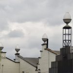 A white-painted warehouse-like building, with decorative eggcups on its roof (one is on some scaffolding above a darkened plate-glass section). There is a tree on the far left.
