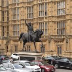 A statue of a man, holding a sword aloft in his right hand and riding a horse, is made of black metal, and stands on a white stone plinth. On the long side the plinth contain a relief, and on the short, by the horse’s front legs, are the words “RICHARD I/COEUR DE LION/1157–1199”. The statue is in a small car park, with low lampposts with glass bulbs around the edge. Behind is the gothic wall of the Houses of Parliament, with narrow notching-arched windows.