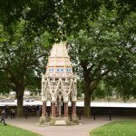 A small building, octagonal in plan, with gothic arches on each side and triangular walls above, with a high pyramidal roof. The arches are made of red stone (bottom) and white stone) above, and the roof is painted in pastel shades in simple geometric patterns, with some dark blue stars near the bottom. The building is in a small park, with a few people walking around, and behind it are trees which largely obscure the view beyond, although the bank of the river and the river itself can be seen below the leaves.