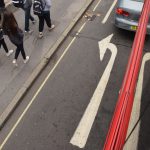 A view down the outside of a bus. The road markings have a large white arrow pointing forward and then curving to the left, which is reflected in the dark bus windows. Part of the red frame of the bus is visible below the windows, and a grey car is stopped just in front of the arrow. On the pavement along side, a group of five people are walking.
