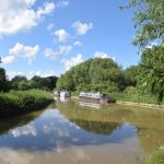 A canal, curving around to the left, with a few narrowboats on it. The towpath, unpaved, is deserted. Trees overhang the canal in the foreground, and line it in the background, and the blue sky with white fluffy clouds is reflected in the murky water.