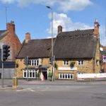 Two Cotswold stone thatched cottages at a traffic-lit road junction (now a restaurant). Brick buildings line the roads from the junction. There are no cars.
