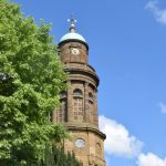 A round stone church tower, getting progressively more recessed as it ascends, with a black clock with gold numerals on the top section. The tower is topped by a copper dome with a weather vane. To the left and at the bottom of the shot are leafy trees.